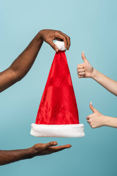 cropped view of interracial couple holding santa hat and showing thumbs up isolated on blue background - Foto, afbeelding