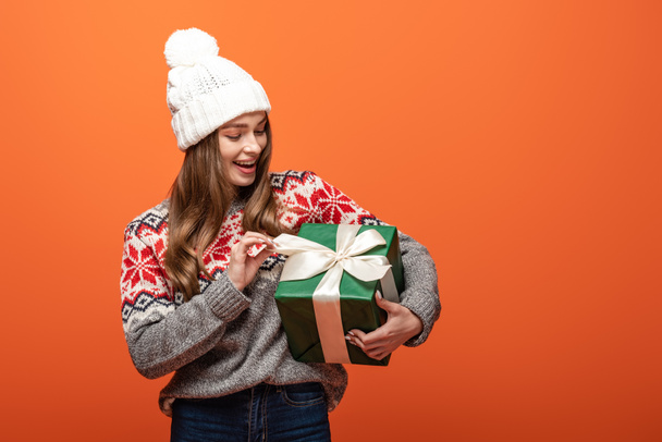 happy girl in winter outfit holding present on orange background - Фото, изображение