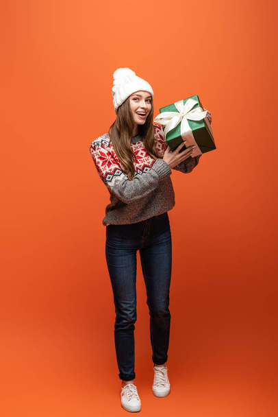 happy girl in winter outfit holding present on orange background - 写真・画像