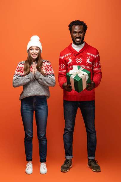 africký Američan muž drží vánoční dárek v blízkosti usmívající se přítelkyně na oranžovém pozadí - Fotografie, Obrázek