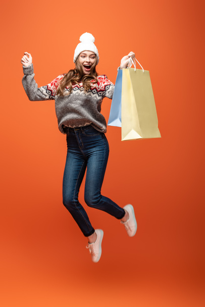 gelukkig meisje in de winter outfit springen met boodschappentassen op oranje achtergrond - Foto, afbeelding