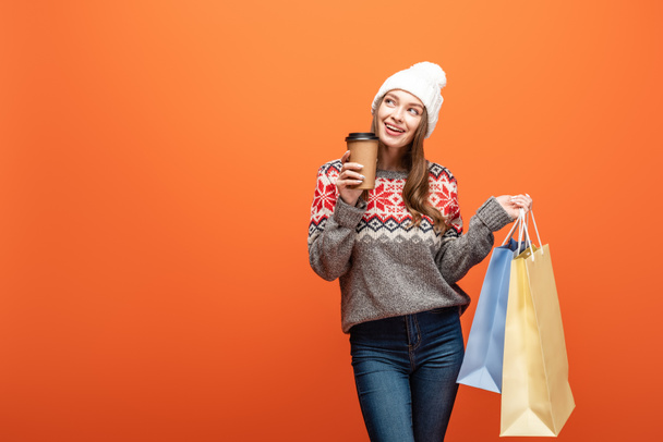 счастливая девушка в зимнем наряде с сумками для покупок и кофе на оранжевом фоне
 - Фото, изображение