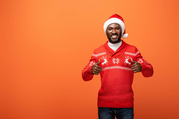 smiling african american man in santa hat and Christmas sweater showing thumbs up on orange background - Zdjęcie, obraz