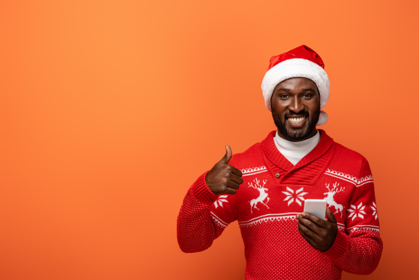 smiling african american man in santa hat and Christmas sweater with smartphone showing thumb up on orange background - Fotografie, Obrázek