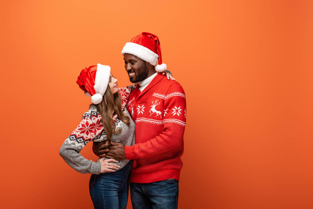 happy interracial couple in santa hats and Christmas sweaters looking at each other and embracing on orange background - Foto, afbeelding
