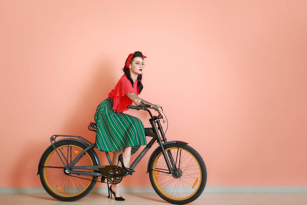 Portrait of beautiful tattooed pin-up woman riding bicycle against color wall - Photo, Image