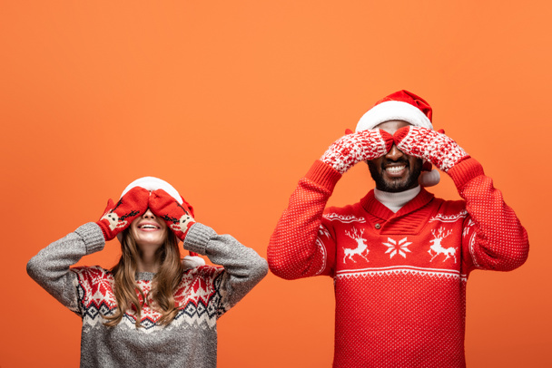 glückliches gemischtrassiges Paar in Weihnachtsmützen, Fäustlingen und Weihnachtspullovern, die die Augen mit Händen auf orangefarbenem Hintergrund bedecken - Foto, Bild