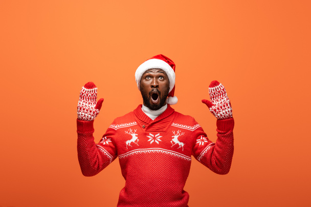 shocked african american man in santa hat, mittens and Christmas sweater on orange background - Fotó, kép