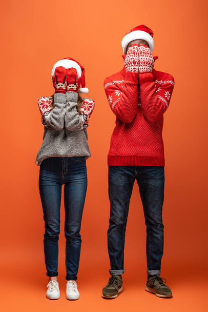 gelukkig interraciaal paar in santa hoeden, wanten en kerst truien bedekken gezicht met handen op oranje achtergrond - Foto, afbeelding