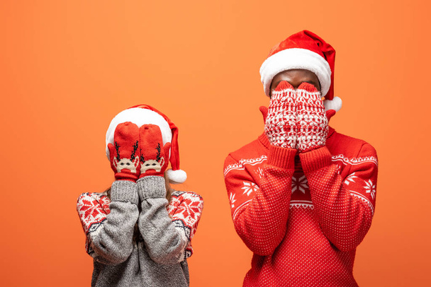 happy interracial couple in santa hats, mittens and Christmas sweaters covering face with hands on orange background - 写真・画像