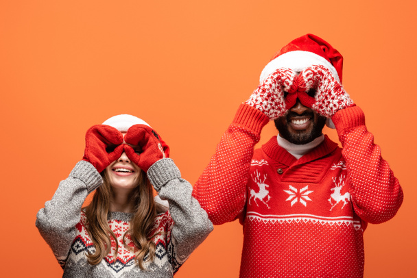 happy interracial couple in santa hats, mittens and Christmas sweaters imitating binoculars with hands on orange background - Photo, Image