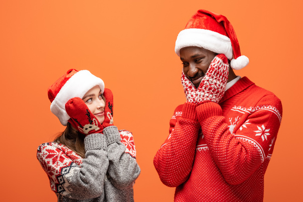 glückliches gemischtrassiges Paar in Weihnachtsmützen, Fäustlingen und Weihnachtspullovern, die Wangen auf orangefarbenem Hintergrund berühren - Foto, Bild