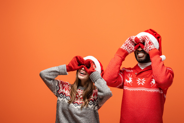 happy interracial couple in santa hats, mittens and Christmas sweaters imitating binoculars with hands on orange background - Fotoğraf, Görsel
