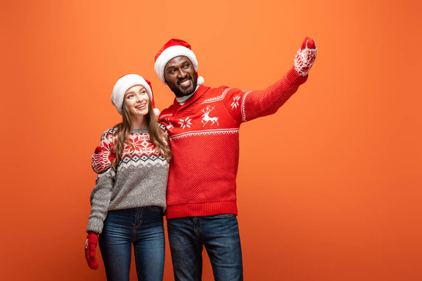 glückliches gemischtrassiges Paar in Weihnachtsmützen, Fäustlingen und Weihnachtspullovern, die sich umarmen und mit der Hand auf orangefarbenem Hintergrund zeigen - Foto, Bild