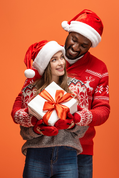 happy interracial couple in santa hats and Christmas sweaters hugging and holding gift on orange background - Photo, Image