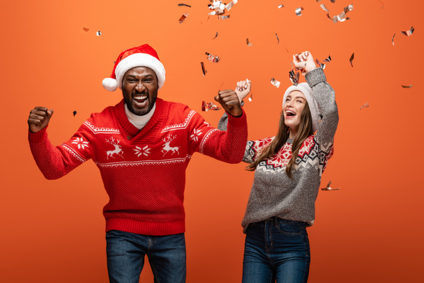 feliz pareja interracial en sombreros de santa y suéteres de Navidad mostrando sí gestos bajo confeti sobre fondo naranja
 - Foto, Imagen