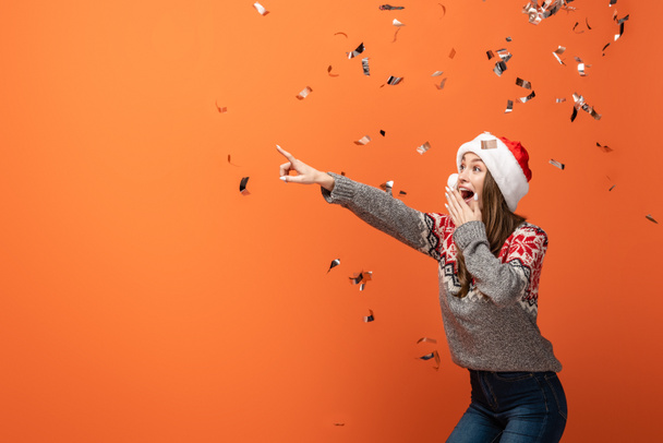 surprised woman in santa hat under falling confetti pointing with finger away on orange background - Photo, Image