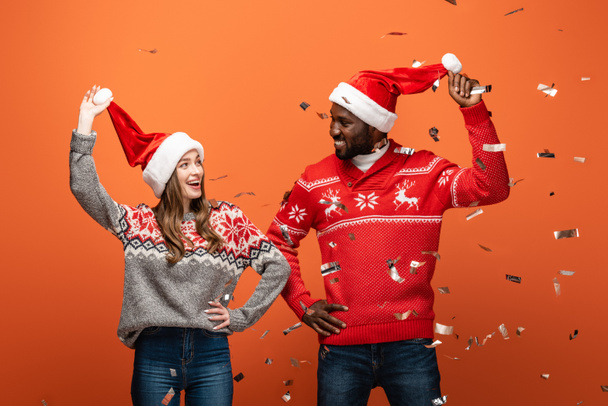 happy interracial couple in santa hats and Christmas sweaters looking at each other under confetti on orange background - Фото, изображение