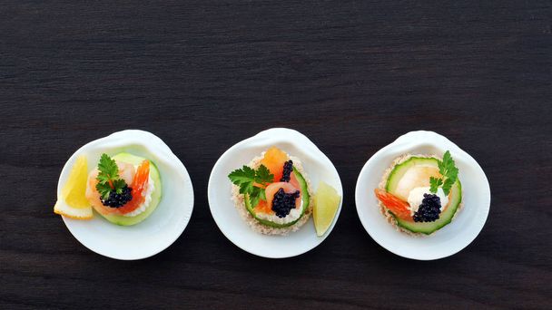 Elegantes canapés con salmón ahumado, camarones y caviar negro en porcelana con forma de concha individual que sirve plato sobre mesa de madera oscura
 - Foto, Imagen