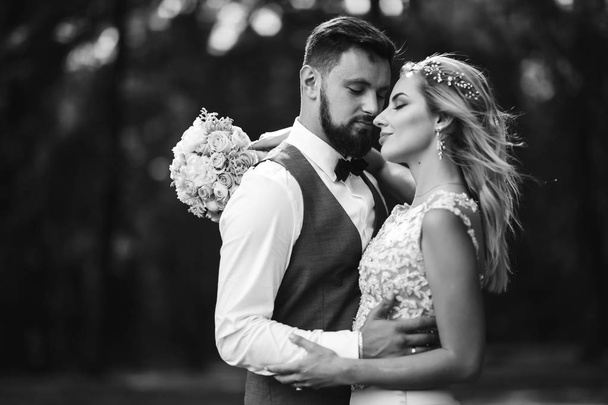 Sensual Black and white foto of bride and groom. Stylish couple of happy newlyweds posing in the park on their wedding day. Handsome bearded groom admires and kisses pretty bride. Together. - Foto, Imagen