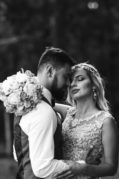 Sensual Black and white foto of bride and groom. Stylish couple of happy newlyweds posing in the park on their wedding day. Handsome bearded groom admires and kisses pretty bride. Together. - Foto, Imagem