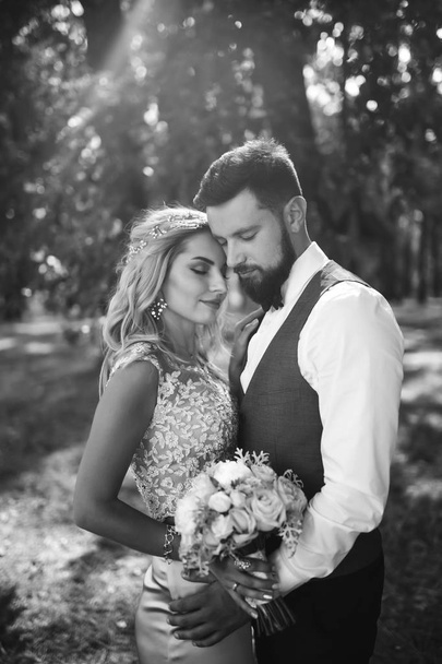 Sensual Black and white foto of bride and groom. Stylish couple of happy newlyweds posing in the park on their wedding day. Handsome bearded groom admires and kisses pretty bride. Together. - Photo, image