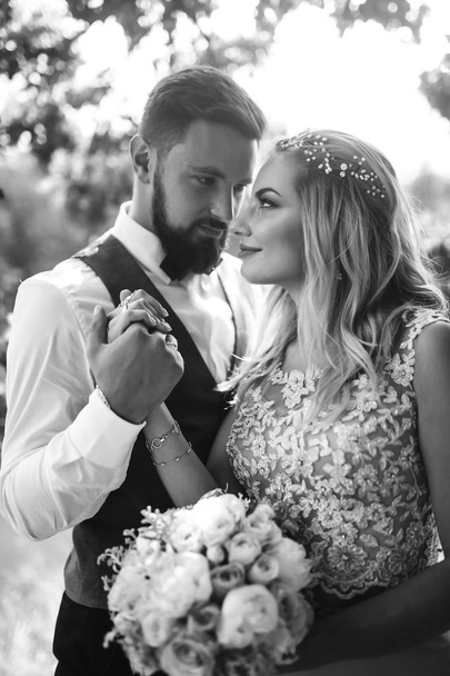 Sensual Black and white foto of bride and groom. Stylish couple of happy newlyweds posing in the park on their wedding day. Handsome bearded groom admires and kisses pretty bride. Together. - Фото, зображення