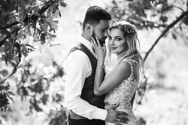 Sensual Black and white foto of bride and groom. Stylish couple of happy newlyweds posing in the park on their wedding day. Handsome bearded groom admires and kisses pretty bride. Together. - Foto, afbeelding