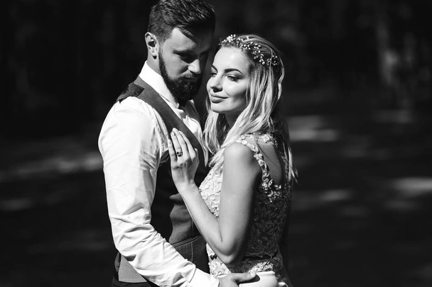 Sensual Black and white foto of bride and groom. Stylish couple of happy newlyweds posing in the park on their wedding day. Handsome bearded groom admires and kisses pretty bride. Together. - Fotografie, Obrázek