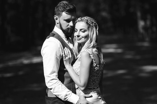 Sensual Black and white foto of bride and groom. Stylish couple of happy newlyweds posing in the park on their wedding day. Handsome bearded groom admires and kisses pretty bride. Together. - Φωτογραφία, εικόνα