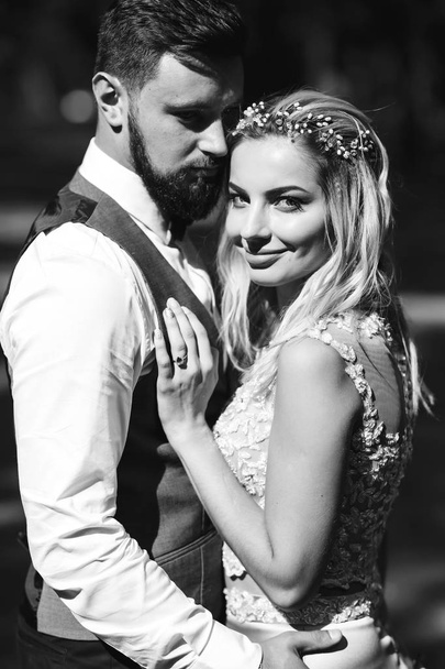 Sensual Black and white foto of bride and groom. Stylish couple of happy newlyweds posing in the park on their wedding day. Handsome bearded groom admires and kisses pretty bride. Together. - Zdjęcie, obraz