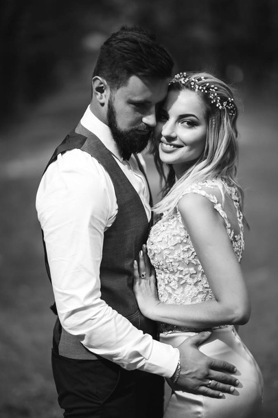 Sensual Black and white foto of bride and groom. Stylish couple of happy newlyweds posing in the park on their wedding day. Handsome bearded groom admires and kisses pretty bride. Together. - Fotó, kép
