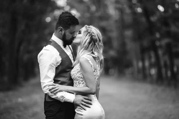 Sensual Black and white foto of bride and groom. Stylish couple of happy newlyweds posing in the park on their wedding day. Handsome bearded groom admires and kisses pretty bride. Together. - Photo, Image
