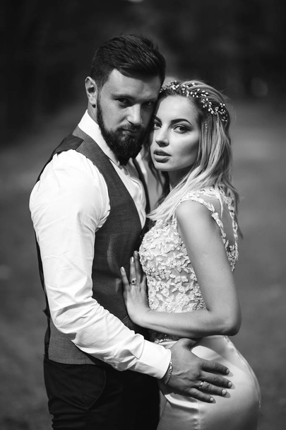 Sensual Black and white foto of bride and groom. Stylish couple of happy newlyweds posing in the park on their wedding day. Handsome bearded groom admires and kisses pretty bride. Together. - Foto, Imagem