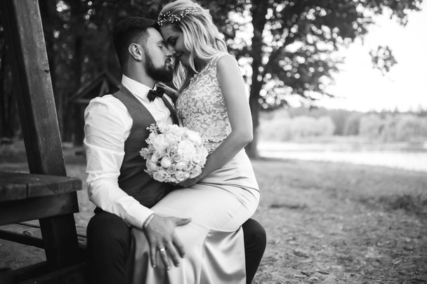 Sensual Black and white foto of bride and groom. Stylish couple of happy newlyweds posing in the park on their wedding day. Handsome bearded groom admires and kisses pretty bride. Together. - Foto, afbeelding