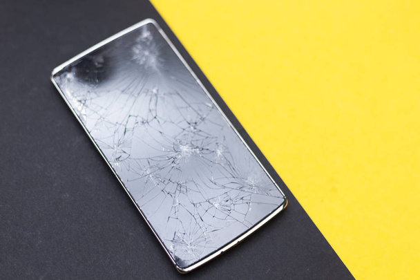 A broken phone on yellow and black background. Crushed device with broken screen representing an accident. Textured screen with damage. Dark glass of a screen. - Valokuva, kuva