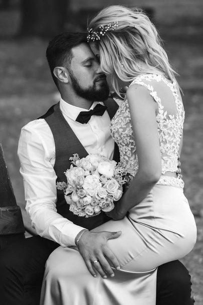 Sensual Black and white foto of bride and groom. Stylish couple of happy newlyweds posing in the park on their wedding day. Handsome bearded groom admires and kisses pretty bride. Together. - Foto, Bild