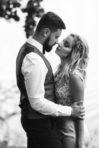 Sensual Black and white foto of bride and groom. Stylish couple of happy newlyweds posing in the park on their wedding day. Handsome bearded groom admires and kisses pretty bride. Together. - 写真・画像