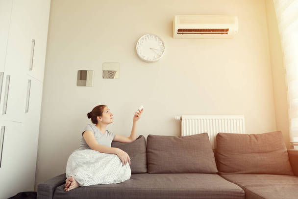 Woman turns on the air conditioner from the remote control sitting on the sofa. - Photo, Image