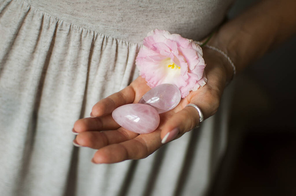 Vrouwelijke handen houden twee Yoni eieren voor vumfit, imbuilding of meditatie zijn gemaakt van roze kwarts en transparante Violet Amethyst met witte bloemen binnenshuis - Foto, afbeelding