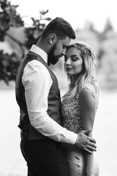 Sensual Black and white foto of bride and groom. Stylish couple of happy newlyweds posing in the park on their wedding day. Handsome bearded groom admires and kisses pretty bride. Together. - Fotoğraf, Görsel