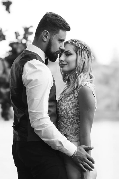 Sensual Black and white foto of bride and groom. Stylish couple of happy newlyweds posing in the park on their wedding day. Handsome bearded groom admires and kisses pretty bride. Together. - Foto, immagini