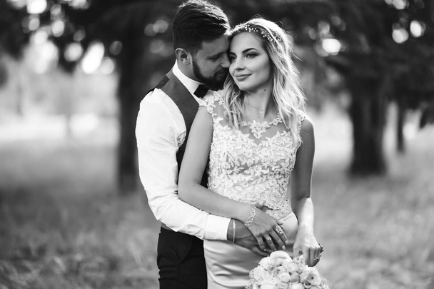 Sensual Black and white foto of bride and groom. Stylish couple of happy newlyweds posing in the park on their wedding day. Handsome bearded groom admires and kisses pretty bride. Together. - Фото, изображение