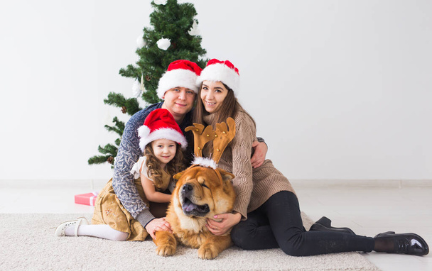 Κατοικίδια ζώα, γιορτές και εορταστική έννοια - Οικογένεια με σκύλο κάθονται στο πάτωμα κοντά στο χριστουγεννιάτικο δέντρο. - Φωτογραφία, εικόνα