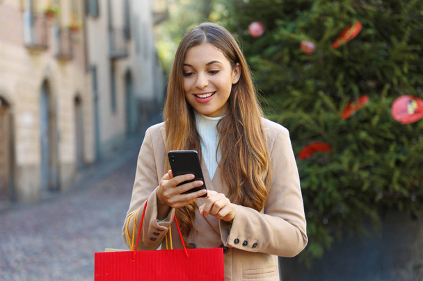Christmas woman buying online on the smart phone in the street with christmas tree on background. - Photo, Image
