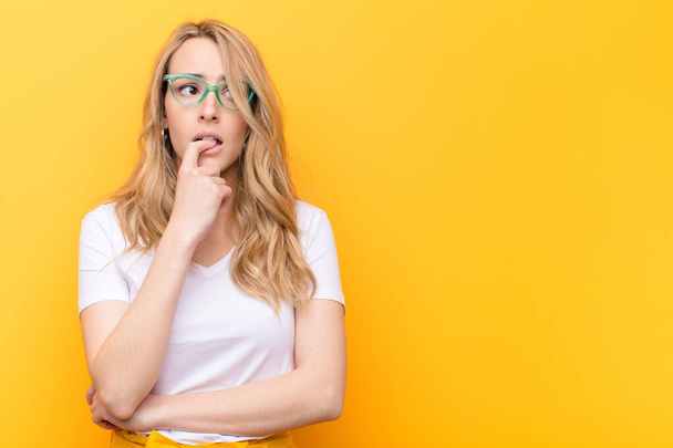 young pretty blonde woman with surprised, nervous, worried or frightened look, looking to the side towards copy space against flat color wall - Foto, Imagen