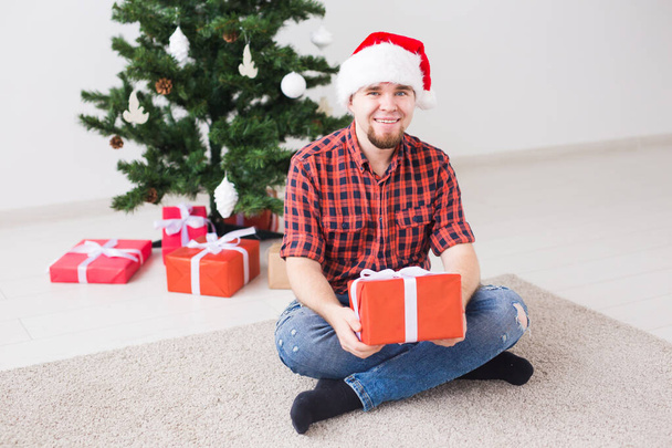 Weihnachts- und Feiertagskonzept - lustiger Mann mit Weihnachtsmütze, der zu Hause im Wohnzimmer ein Geschenk hält - Foto, Bild