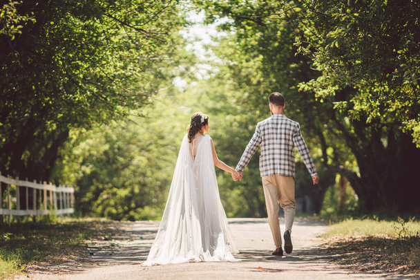 Happy bride and groom holding hands and walking in garden wedding day. Back view of charming stylish newlyweds holding hands while walking in park forest. Path to future life. happy marriage moments - Foto, immagini