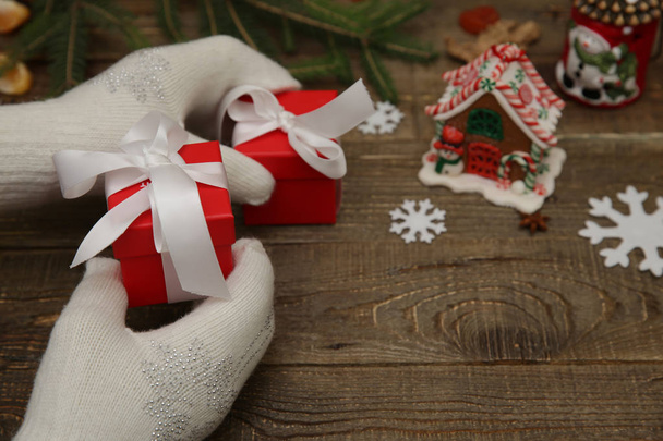 Руки Санта Клауса, чоловіки або жінки в білих рукавицях, тримають дві червоні подарункові коробки з білим луком, на задньому плані різдвяних кульок і ялинок на дерев'яному столі. Поняття Різдва і Нового року. - Фото, зображення