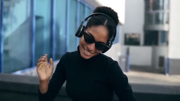 Pretty afro girl happy smiling. Stunning african woman with light-brown skin relaxing in headphones and funny dancing on modern city background. Wearing black sweater and stylish black sunglasses - 映像、動画